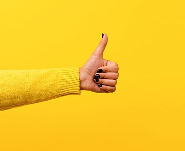 Ein ausgestreckter Arm der von links ins Bild ragt und den Daumen hoch zeigt, vor gelbem Hintergrund mit gelbem Longsleeve.