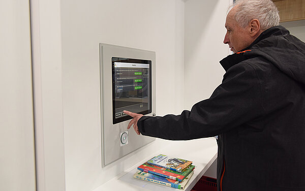 Ein Mann steht an einem Ausleihautomaten und bedient jenen. Vor ihm liegt ein Stapel Bücher.