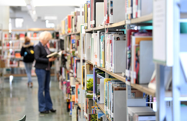 Eine Erwachsene am Bücherregal. Das regal ist im Vordergrund scharf, die Person im Hintergrund unscharf