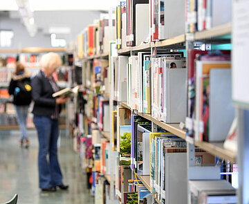 Eine Erwachsene am Bücherregal. Das regal ist im Vordergrund scharf, die Person im Hintergrund unscharf