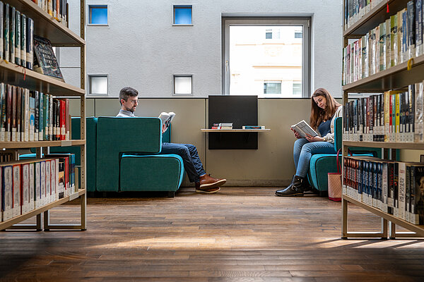 Zwei Menschen sitzen sich vor Rücherregalen gegenüber und lesen.