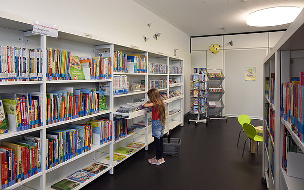 Ein Mädchen steht vor dem großen Regal mit Kinderbüchern und stöbert.