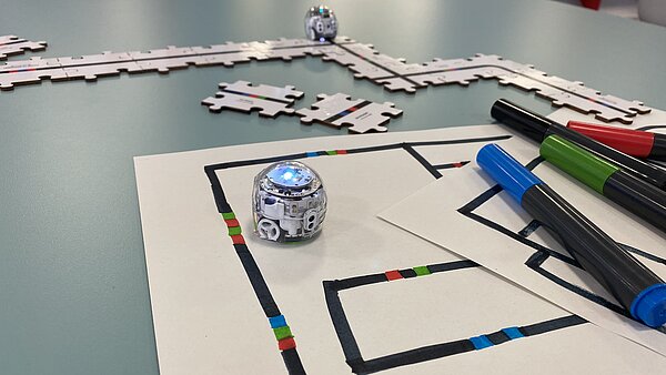 Eine Nahaufnahme der kleinen, runden Ozobot-Roboter, die mit Farbcodes programmiert werden können. Der Roboter im Vordergrund steht auf Papier, das mit Farbcodes bemalt ist, nebendran liegen Filzstifte und im Hintergrund ist ein Ozobot auf Puzzleteilen zu sehen, auf denen Farbcodes gedruckt sind.