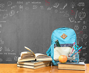 Auf einem Tisch vor einer grauen Tafel stehen ein blauer Schulrucksack, gestapelte, teilweise aufgeschlagene Bücher, ein befüllter Stiftebecher und ein Apfel.