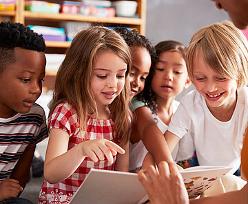 Eine Gruppe kleiner Kinder sitzt auf dem Boden, während ihnen ein Buch von einer Frau vorgelesen wird. Sie schauen fröhlich auf das Buch.