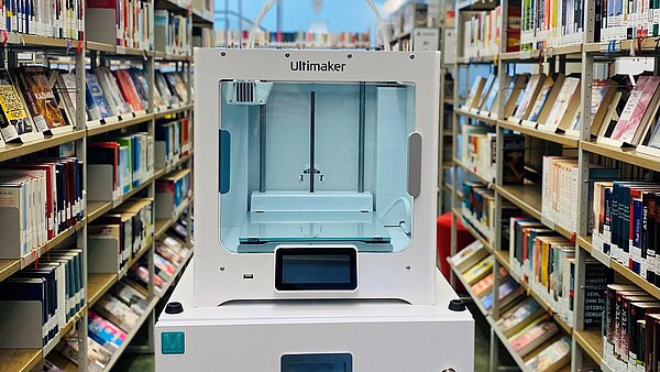 Der 3D-Drucker steht inmitten der Bibliothek zwischen Bücherregalen