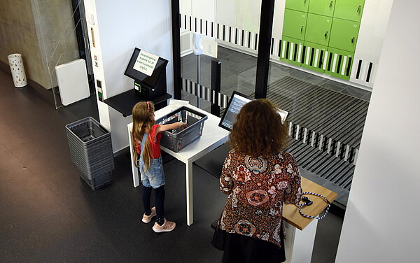 Ein Mädchen steht an den Ausleihautomaten und leiht Bücher aus, die sie aus ihrem Korb holt. Neben ihr steht eine Mitarbeiterin die ihr dabei hilft