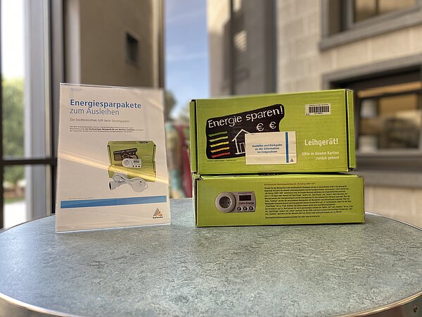 Zwei Energiesparpakete liegen gestapelt auf einem Tisch. Daneben der Aufsteller mit der Information zu den Energiemessgeräten, die man ausleihen kann.