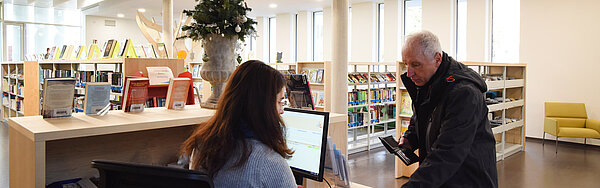 Ein Mann steht an der Ausleihtheke. Eine Mitarbeiterin leiht ihm eine Tonie-Box aus. Im Hintergrund ist die restliche Bibliothek zu sehen mit vielen Bücherregalen.