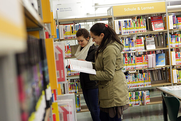 Zwei Mädchen stehen am Bücherregal in der Abteilung "Schule und Lernen" im Neuen Ständehaus und gucken sich lachend ein Buch an.
