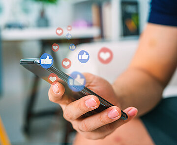 Eine Nahaufnahme von einer Hand die ein Smartphone hält, darüber steigen kleine Like-Daumen und Herzen in die Luft 