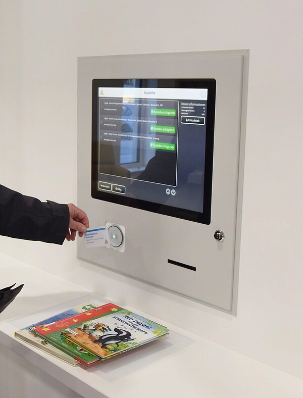 Ein Foto von unserem Ausleihatuomaten, der in der Wand verbaut ist. Man sieht einen Bildschirm, der drei erfolgreich verbuchte Medien anzeigt. Eine Person hält ihren Bibliotheksausweis vor die Schaltfläche des Automaten. Vor dem Automaten liegen drei Bücher auf der Ausleihplatte.