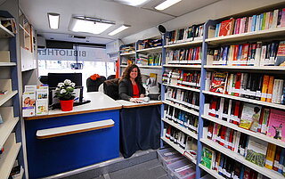 Eine Mitarbeiterin der Bibliothek sitzt im Medienbus an der Ausleihtheke und lächelt in die Kamera. Drumherum volle Regale