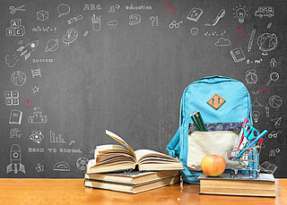 Auf einem Tisch vor einer grauen Tafel stehen ein blauer Schulrucksack, gestapelte, teilweise aufgeschlagene Bücher, ein befüllter Stiftebecher und ein Apfel.