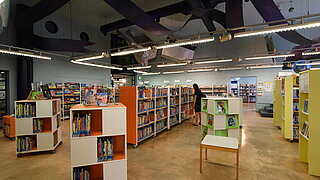 Der Innenraum der Bibliothek mit vielen gefüllten Bücherregalen und einer Frau, die an einem Regal stöbert.