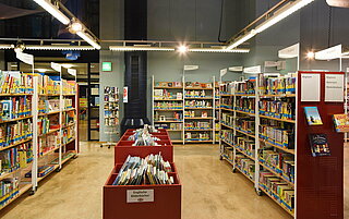 Volle Bücherregale säumen den linken und rechten Bildrand. In der Mitte stehen rote Tröge in denen sich Bilderbücher in vielen Sprachen der Welt befinden