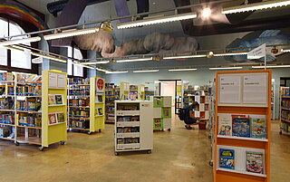 Die Kinder- und Jugendbibliothek von innen. Viele bunte Bücherregale sind zu sehen. Eine Frau sitzt in der Hocke vor einem davon und stöbert