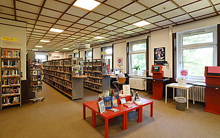 Die Räumlichkeiten der Amerikanischen Bibliothek von innen. Viele Bücheregale und Tische mit Büchern drauf. Im Hintergrund sitzt ein Mann am Computer.