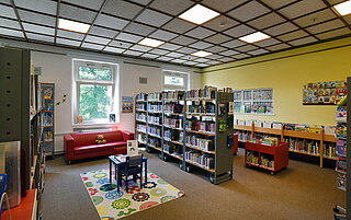Der Kinderbereich in der Amerikanischen Bibliothek. In der Mitte steht ein Tisch mit Stühlen auf einem bunten Teppich. Im Hintergrund Bücherregale und ein rotes Sofa auf dem ein Teddybär sitzt.