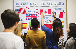 Jugendliche stehen mit dem Rücken zur Kamera gewandt vor einem Whiteboard an dem verschiedenen Landesflaggen hängen (USA, Großbritannien, Kanada, Frankreich, Spanien), oben drüber steht "If you want to say hello" und über den jeweiligen Flaggen auf Landessprache das Wort Hallo