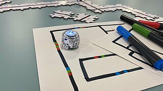 Eine Nahaufnahme der kleinen, runden Ozobot-Roboter, die mit Farbcodes programmiert werden können. Der Roboter im Vordergrund steht auf Papier, das mit Farbcodes bemalt ist, nebendran liegen Filzstifte und im Hintergrund ist ein Ozobot auf Puzzleteilen zu sehen, auf denen Farbcodes gedruckt sind.