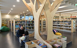 Der Innenraum unserer Zweigstelle Mühlburg. Dort steht ein großes Sitzelement auf dem ein Erwachsener mit einem kleinen Kind sitzt und sich ein Buch anschaut. Im Hintergrund sind volle Bücherregale zu sehen.