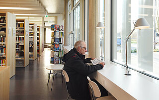 Ein Mann sitzt mit einem aufgeschlagenen Buch in den Händen an einem Tisch vor großen Fenstern. Im Hintergrund befinden sich volle Bücherregale.