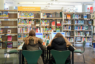 Zwei Mädchen sitzen am Tisch in der Abteilung "Schule und Lernen" im Neuen Ständehaus an Recherchecomputern mit dem Rücken zur Kamera. Im Hintergrund sind Bücherregale zu sehen.