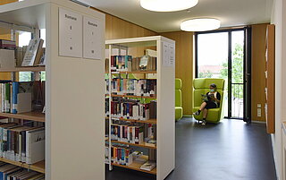Ein weiterer Raum in der oberen Etage ist abgebildet. Hier sind wieder zwei Regale mit Büchern zu sehen und im Hintergrund ein großer grüner Sessel vor einem großen Fenster. Auf dem Sessel sitzt eine Leserin