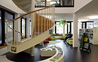 Der Innenraum der Stadtteilbibliothek Grötzingen. Ein heller, moderner, großer Raum. Mittig findet sich eine Wendeltreppe in die obere Etage. Im Hintergrund ist der Kinderbereich mit Marienkäfer-Sitzkissen und einer grünen Sitzbank zu erahnen.