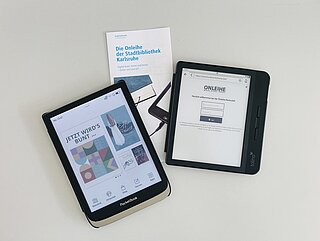Ein eingeschaltetes Pocketbook, ein Tolino, der die Anmeldemaske der Onleihe zeigt, und ein Flyer mit der Aufschrift "Die Onleihe der Stadtbibliothek Karlsruhe". Alles auf einem hellen Tisch liegend.