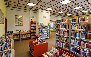 Noch ein Bild aus der Kinderabteilung der Amerikanischen Bibliothek. Viele Bücherregale sind zu sehen und in der Mitte rote große Tröge mit Bilderbüchern drin.