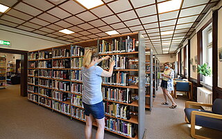 Eine Frau steht vor einem großen Bücherregal und scheint ein Buch zu suchen. Weiter hinten im Raum steht eine weitere Frau und stöbert in den gefüllten Bücherregalen.