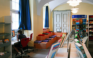 Blick in die Stadtteilbibliothek Durlach. Ein Junge sitzt am PC. Im Hintergrund ist die Treppe mit bunten Sitzkissen zu sehen, rechts Bücherregale