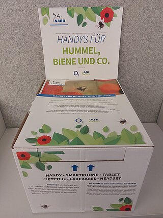 Die Altgeräte-Recycling-Box ist eine weiße Pappbox mit der Aufschrift "Handys für Hummel, Biene und Co.", dem NABU-Logo und weiteren Informationen