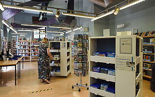 Hier sind die kastenartigen Türme im Bild, in denen Konsolenspiele und DVDs liegen. Eine Mitarbeiterin steht an einem dieser speziellen Regale und räumt etwas ein. Im Hintergrund weitere Bücherregale 