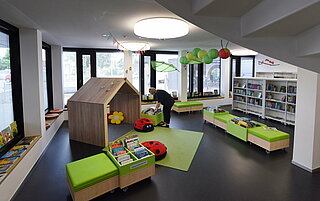 Im Kinderbereich steht ein Holzhaus in dem man sitzen kann. Es finden sich eine grüne Sitzbank und grüne Sitzwürfel sowie ein grüner Teppich vorm Häuschen und Marienkäfer-Sitzkissen auf dem Teppich. Im Hintergrund eine große Fensterfront und Bücherregale