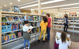 Vor einem Bücherregal stehen zwei Kinder, sie schauen sich Bücher an. Eine Mitarbeiterin erklärt zeigt ihnen Bücher. Ein drittes Kind sitzt und schaut sich ein Buch an.