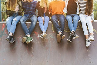 Sechs Jugendliche sitzen nebeneinander auf einer Mauer, von hinten scheint die Sonne auf sie, sie sind ohne Köpfe zusehen