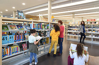 Kinder stehen an einem Bücherregal. Ein Kind schaut ein Buch an, ein anderes Kind redet mit einer Mitarbeiterin, ein drittes Kind sitzt und liest ein Buch. Im Hintergrund läuft eine Frau durchs Bild und lacht den Kindern zu.