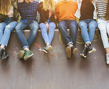 Sechs Jugendliche sitzen nebeneinander auf einer Mauer, von hinten scheint die Sonne auf sie, sie sind ohne Köpfe zusehen