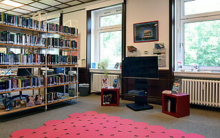 Auf dem Bild ist links ein gefülltes Bücherregal angeschnitten. Daneben erkennt man eine Spielekonsole und einen Bildschirm. Auf dem Boden liegt roter Teppich.