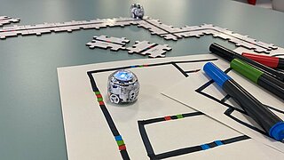 Eine Nahaufnahme der kleinen, runden Ozobot-Roboter, die mit Farbcodes programmiert werden können. Der Roboter im Vordergrund steht auf Papier, das mit Farbcodes bemalt ist, nebendran liegen Filzstifte und im Hintergrund ist ein Ozobot auf Puzzleteilen zu sehen, auf denen Farbcodes gedruckt sind.