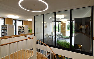 Hier ist die obere Etage abgebildet. Hinter einer großen Glasfront findet sich das Lernstudio. Auf der linken Seite sind Bücherregale zu erkennen
