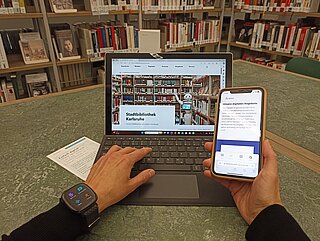 Eine Person sitzt vor aufgeklapptem Laptop, man sieht die Hände angeschnitten am Laptop, am linken Handgelenk trägt sie eine Smartwatch, in der rechten Hand hält sie ein Smartphone. Auf den Geräten ist die Stadtbibliothek Homepage geöffnet. Im Hintergrund ist ein Bücherregal zu sehen.