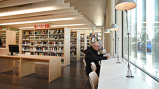 Ein Mann sitzt mit einem Buch an einem hellen Tisch vor einer großen Fensterfront. Im Hintergrund ist die restliche Bibliothek zu sehen mit vielen Bücherregalen