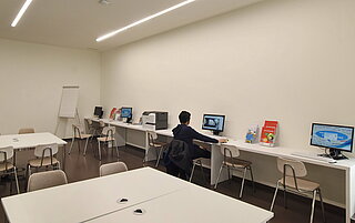 Das Lernstudio in Mühlburg ist ein großer heller Raum mit Tischgruppen an denen Stühle stehen. Hinten an der Wand sind Tische nebeneinander aufgereiht. Auf ihnen stehen PCs. An einem PC sitzt ein junger Mann.