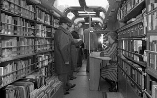 Die erste Generation des Medienbusses von innen. Auf dem schwarz-weiß Bild stehen Menschen vor den Regalen im Bus