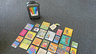 Eine Auswahl der Bücher liegen auf dem Boden und daneben steht der Regenbogenkinderbücherkoffer.