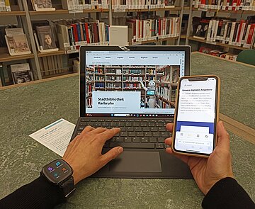Eine Person sitzt vor aufgeklapptem Laptop, man sieht die Hände angeschnitten am Laptop, am linken Handgelenk trägt sie eine Smartwatch, in der rechten Hand hält sie ein Smartphone. Auf den Geräten ist die Stadtbibliothek Homepage geöffnet. Im Hintergrund ist ein Bücherregal zu sehen.